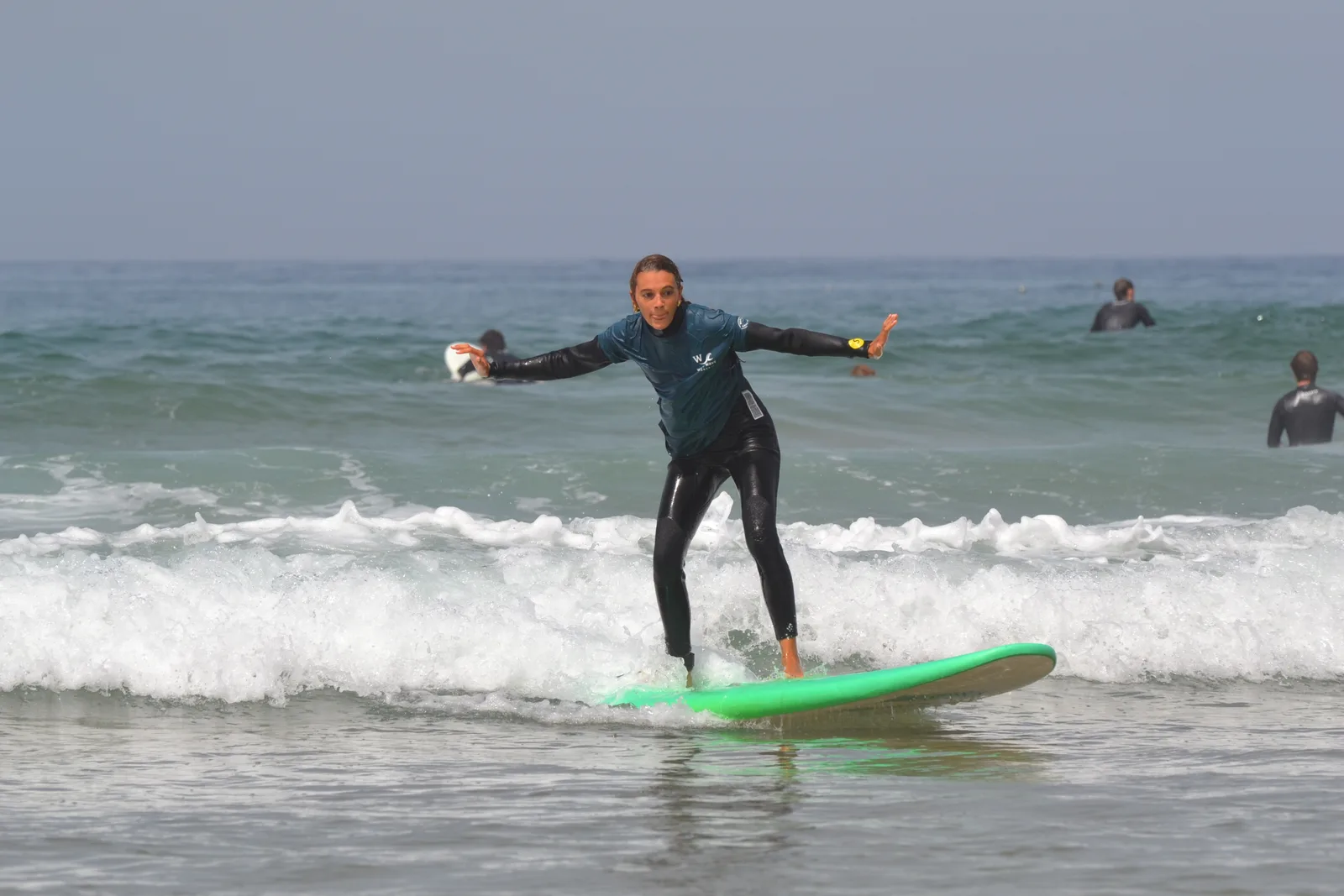 A surfer balancing on a green board in whitewater, with other surfers in the background.